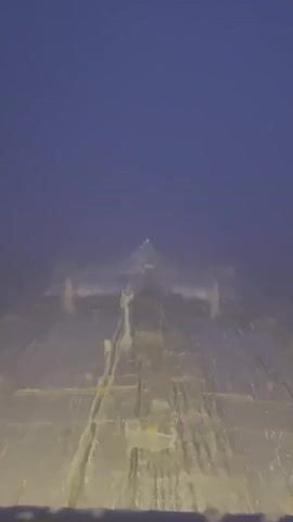 The video shows an underwater view looking forward from a vehicle. The water is murky and a deep blue. Below the water's surface, a large, pale, metallic structure is visible. It appears to be a ship or some type of marine vessel, with a long, flat deck and a central superstructure with multiple levels. There are railings and pipelines visible on the deck, as well as some scattered debris. The vehicle is moving slowly, and the underwater environment is lit by artificial lights from the vehicle, creating a somewhat surreal and ghostly effect.