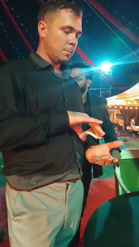 A man is performing a card trick outdoors at night. He stands near a green table with a glass of red wine on it. He wears a black button-down shirt and gray pants. He shuffles a deck of cards, then places them face down on the table. Behind him, other people are gathered, some seated under a large tent decorated with string lights. A metal structure stands behind the tent. Red banners hang overhead.
