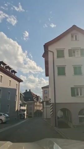 A first-person view from a vehicle driving down a narrow street lined with buildings. The sky is blue with scattered white clouds. On the left, there's a light-colored building with several windows. Further down the street on the left, there are buildings with darker roofs and more windows. On the right, a large, light-colored building dominates the view, with green shutters visible on some windows. The street is paved, and a few vehicles are visible parked along the side. The video captures the movement of the vehicle as it proceeds through the street.