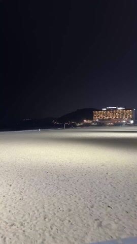 A video shows a sandy beach at night. Initially, the beach is brightly lit with a large building visible in the background. The video then moves toward the city, revealing a pier and several skyscrapers lit up in the distance. People can be seen walking along the beach. The sky is black.
