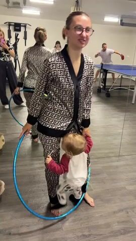 The video shows a woman in pajamas holding a hula hoop around her waist and interacting with a toddler who is also inside the hoop. In the background, a man is playing table tennis. The scene appears to be indoors, possibly in a gym or recreational area, with mirrors reflecting the activity.