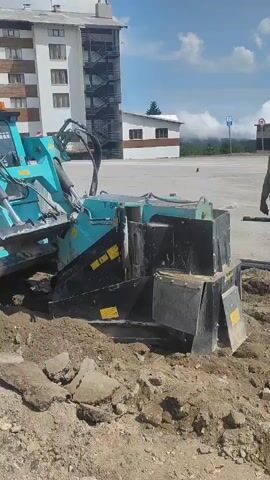 A turquoise and black trenching machine with rotating cutting blades is digging a trench in a dirt and asphalt area. The machine has tracks and a wheeled front end. It is positioned with its cutting head lowered into the ground, excavating material. Chunks of asphalt and dirt are visible around the trench. In the background, there is a building with multiple floors and balconies, and a blue sky with some clouds. A person wearing a high-visibility vest is standing near the machine, observing its operation.