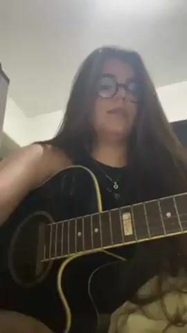 A woman with long brown hair and glasses is sitting and playing a black acoustic guitar. She is singing while playing the guitar. She's wearing a black tank top and a necklace.  She is indoors, possibly in a bedroom, with a light-colored wall behind her.  There is a door visible in the background.