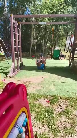 A child is swinging upside down on a swing set in a backyard playground. The child is wearing a blue shirt and dark shorts. The swing set has a wooden frame with a ladder and two swings. In the background, there is a green slide and another person on the playground. The ground is covered with green artificial turf and dirt. A red plastic toy is in the foreground. The trees in the background suggest it is a sunny day.