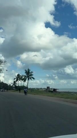 The camera, likely inside a vehicle, films a road next to an ocean coastline. Palm trees line the road. People can be seen walking and riding motorbikes.  Buildings are visible in the distance. White, puffy clouds dominate the blue sky.
