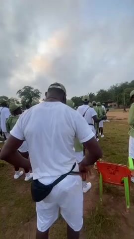 A group of people, mostly young adults, are gathered in an outdoor grassy area, likely a field or parade ground, at what appears to be Emure-Ekiti, Nigeria. The subjects are predominantly wearing matching uniforms: light green shirts, white shorts, and green caps. Some are holding musical instruments, specifically drums and what appear to be drumsticks. One individual in the foreground, facing away from the camera, wears a white t-shirt and white shorts, with a black fanny pack around their waist. This person is holding drumsticks, suggesting they are participating in or leading a drum ensemble.

The overall activity seems to be a practice session or preparation for a performance, likely a parade or ceremony, given the uniform attire and musical instruments. The time of day is early morning or late afternoon, as indicated by the soft, diffused light and overcast sky. The weather appears to be cloudy, possibly humid.

In the foreground, an orange and green plastic chair is visible. Some of the individuals are holding up their drumsticks, creating a sense of anticipation or instruction. The atmosphere appears orderly and focused. A few people are standing in the background, observing or waiting their turn. The scene is set against a backdrop of trees and shrubs, with a hint of buildings in the distance, common in rural or semi-rural Nigerian settings.

There is some visible text on the orange chair, but it is difficult to decipher clearly. The focus of the image is the group and their synchronized activity, suggesting a communal event or training exercise.