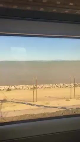 The video shows a view from inside a moving train. The train is passing near the ocean. The camera pans from left to right across the inside of the train. There are several rows of purple and grey seats. The train appears empty. The view outside the window includes water, a beach with a few cars, and a highway. There is a no smoking sign on the wall.