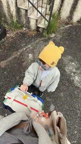 The image presents an overhead view of an outdoor scene in Nerin, Montenegro, featuring a young child and portions of an adult.

In the center, a young child is seen crouching or sitting on a dark, pebbled asphalt surface. The child wears a distinctive bright yellow beanie with two small, rounded ear-like elements, light blue-framed glasses, a light grey bomber jacket, and dark trousers with dark shoes. The child is focused on a cream-colored bag with colorful striped accents in red, green, and blue, and a black handle. Their right hand holds a small, dark object, possibly a remote or device, touching the bag.

In the immediate foreground, the lap and upper legs of an adult, clad in grey shorts, are visible. They hold a white tote bag with brown handles and a fluffy white interior lining. Inside this bag, a small, orange-and-white stuffed animal or toy is nestled, appearing to be connected to blue tubing or straps.

The setting is an outdoor area, likely a paved pathway or a backyard. To the left, a dark car tire is partially visible. In the background, a set of weathered concrete stairs with a black metal railing leads upwards. Sparse green vegetation grows through cracks in the ground and along the base of the stairs, suggesting a somewhat untended environment. The lighting is natural and even, indicative of daytime, possibly under an overcast sky. No discernible text is visible on the subjects' clothing or the bags. The scene depicts a quiet moment, with the child engaged with their belongings.