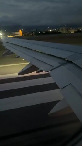 The video shows the wing of an airplane on a runway at dusk. The runway lights are visible, and there is a city skyline in the distance. The wing is large and white, with a purple and pink logo on the tip. The engines are not visible, but the wing appears to be attached to a fuselage. The plane is stationary on the runway. The sky is dark and cloudy.