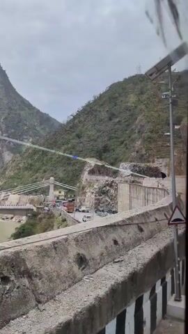 The video pans across a mountainous landscape with a river flowing through a valley. A concrete barrier is in the foreground, obscuring the bottom portion of the scene. In the middle ground, a long suspension bridge spans the river, with traffic visible on its deck. Several vehicles, including trucks and cars, are on the bridge and on the road leading up to it. The mountains on either side of the valley are steep and covered in vegetation, with some rocky outcrops visible. There are some signs of human construction, including retaining walls and roads carved into the mountainside. The sky is overcast.