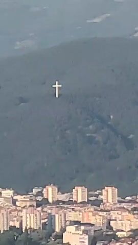 The video presents a panoramic view of a city and its surrounding landscape, as seen from an elevated position. The central feature is a large white cross prominently situated on a distant, tree-covered mountain.

The video opens with a close-up shot (0:00-0:02) of the white cross against the dark green mountain, with the upper parts of multi-story city buildings visible below. The sky appears hazy. The camera then zooms out slightly (0:02-0:03), revealing more of the city nestled in a valley, flanked by green hills and fields. The cross remains a focal point on the mountain.

The final shot (0:03-0:05) provides a wider perspective, showing the vastness of the valley and the city stretching across it. Lush green trees and bushes fill the foreground, framing the expansive view. The white cross is now a smaller, distinct mark on the distant mountain. The sky in this wider shot is partly cloudy with patches of blue.

The scene depicts a blend of urban and natural environments, with the cross serving as a notable landmark overlooking the city. The time of day appears to be daytime, with varying sky conditions from hazy to partly cloudy. No people or text are visible in the video, which is set in Selishte, Bulgaria.