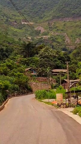 The video shows a view of a winding road in a mountainous, tropical-looking area. The road is asphalt and is lined on one side by a low stone wall and lush green vegetation. Across the road, there is also dense vegetation. The camera appears to be moving along the road, capturing the scenery. Further up the mountain, a building is visible nestled among the trees. As the video progresses, a person riding a motorbike comes into view and passes by. The overall environment is lush and green, suggesting a humid climate.