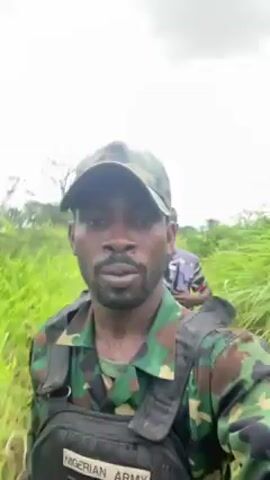 A man in a Nigerian Army uniform and a baseball cap walks through tall grass. He has a rifle slung over his shoulder and a flak jacket labeled "NIGERIAN ARMY." Another man walks behind him, also carrying a rifle. They are surrounded by green vegetation. The sky is overcast. The man in the foreground is filming himself.