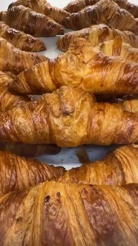 The media file showcases a close-up, overhead view of a large batch of freshly baked croissants, meticulously arranged in rows. The croissants are golden brown and appear flaky and buttery, with visible layers of dough. They are stacked on what appears to be a white surface, possibly a baking tray or a display counter. The lighting suggests a well-lit indoor environment, possibly a bakery or pastry shop. The focus is entirely on the croissants, highlighting their texture and appetizing appearance. No people, text, or specific location cues within Adeje, Spain are visible in the frame. The overall impression is one of abundance and the enticing presentation of baked goods.