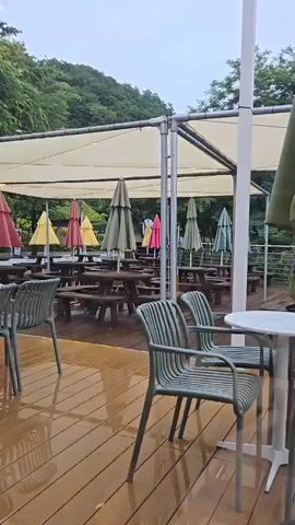 The video shows an outdoor seating area, possibly a cafe or restaurant, with several picnic tables and chairs under a large white awning. The ground, made of wooden planks, is wet and reflects the surrounding light, suggesting it has been raining or is currently raining. Colorful umbrellas are set up over the tables, in shades of red, yellow, and green. In the background, a line of trees and a grassy slope are visible, indicating a natural or park-like setting. Two people are faintly visible in the background, one walking near the tables and another standing further back. A large, closed pink umbrella is prominently featured in the foreground, to the left, mounted on a heavy black base. Several green chairs are arranged around white tables in the foreground. The overall atmosphere appears calm and perhaps a bit deserted, possibly due to the weather.