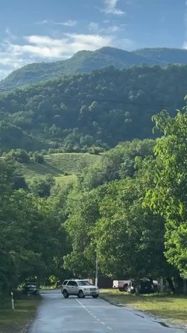 This video shows a road lined with trees on either side. Mountains covered in lush green foliage rise in the background, with a clear blue sky above. Several cars are parked along the right side of the road. The road itself is paved and appears wet, suggesting recent rain. The overall scene is a tranquil, natural landscape.