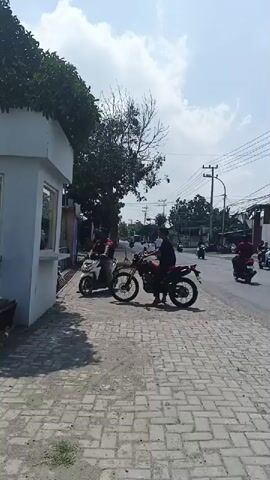 The video shows a bright, sunny day with a clear sky. The scene appears to be an outdoor area with a paved walkway and a road. On the left side of the frame, there is a small white booth with a window. A person is visible inside the booth. In front of the booth, two motorcycles are parked, one white and one red. Two individuals are interacting with the motorcycles. One person is standing next to the white motorcycle, and another person is seated on the red motorcycle. In the background, there is a tree with green foliage and a bare, leafless tree. Further in the background, there are buildings, power lines, and other vehicles on the road, indicating a developed area. The overall atmosphere is casual and everyday.