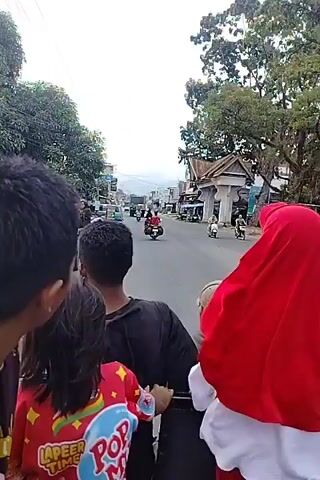 The video shows a view from the back of a moving vehicle, possibly a motorcycle or a small truck. In the foreground, several people are visible from behind, including a person wearing a red headscarf. The road ahead is lined with buildings that appear to be shops or houses. There are other vehicles on the road, including motorcycles and cars. The sky is cloudy. The overall impression is a glimpse of everyday life on a street in a populated area.