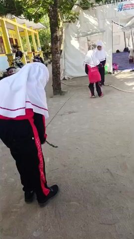 The video captures an outdoor scene in Simpangjaya, Indonesia, featuring three young females engaged in or observing a jump rope game under bright sunlight.

In the foreground, a girl stands with her back to the camera, wearing a white hijab with a red trim, a black long-sleeved top, and black athletic pants with red stripes down the sides, which appear to have white text (possibly a brand name like 'ADIDAS'). She is wearing black shoes and holds a small, green, rope-like object in her right hand, suggesting she is waiting her turn or preparing to play.

Further in the background, two other girls are actively involved in jumping rope. One girl, dressed in a white hijab, a red top, and black pants, is shown mid-jump over the rope. Another girl, also in a white hijab and dark clothing, is turning one end of the rope.

The setting appears to be a paved or dirt area, possibly a schoolyard or community space. To the left, a yellow building is visible with parked motorcycles. A tree stands in the mid-ground. To the right, a large white tent structure is set up, with a blue tarp or mat visible near its entrance, hinting at a temporary shelter or event area.

The bright daylight casts clear shadows, indicating a sunny day. In the top right corner of the frame, a logo with the partially visible Indonesian text "PENDIDIKAN BERMUTU UNTUK SE..." (likely "Pendidikan Bermutu untuk Semua," meaning "Quality Education for All") is present, reinforcing the potential educational context of the location.