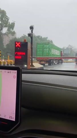 The video captures a view from inside a vehicle, looking through the windshield at a toll booth in what appears to be a rainy or misty environment. The weather is overcast and the road surface is wet, with visible water splashes.

In the foreground, a black car dashboard is visible, with a large touchscreen display on the left. The display shows a navigation interface with a map and a time of 11:58. The temperature outside is indicated as 25.0.

The main focus of the scene is a toll booth. A red "X" is illuminated on a digital sign, indicating that the lane is closed or there is an issue. Below the "X", Chinese characters are displayed. The text visible reads "广东换乘站" (Guangdong Transfer Station), "收款0.00元" (Collection 0.00 Yuan), and "付费" (Payment). A yellow barrier arm is extended across the lane, blocking traffic.

Several vehicles are visible in the background. A large green shipping container truck with "EVERGREEN" branding is stopped at or approaching the toll booth. Other trucks and cars are also present, suggesting moderate traffic. The overall setting appears to be a highway or a major road in a suburban or semi-urban area, with trees and buildings visible in the distance. The time of day appears to be late morning. The atmosphere is somewhat subdued due to the weather conditions.