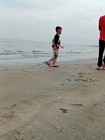 The video presents a peaceful scene on a sandy beach in Blanggeulumpang, Indonesia, under an overcast sky. Three individuals are visible on the shore. A young child, dressed in a black t-shirt and light-colored pants with sandals, walks slowly towards the ocean, his back to the camera. To his right, an older male figure in a red t-shirt and dark pants stands, also facing the sea, observing the water. Further to the right and slightly behind him, another person is seen crouching down on the sand.

The beach is characterized by light grey-brown sand, which appears damp in areas and has some scattered dark patches or debris. Gentle waves lap at the shore, and the calm ocean stretches into the distance, meeting a hazy horizon. The sky is uniformly cloudy, diffusing the light and suggesting an overcast day, possibly early morning or late afternoon. Distant vegetation or land is faintly visible along the coastline on the right side of the frame. The scene conveys a quiet moment of relaxation by the sea. No text is visible in the video.