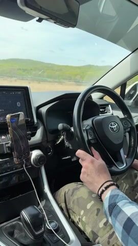 Here's a description of the video:

A person is driving a Toyota RAV4. The view is from inside the car, showing the driver's hands on the steering wheel, the dashboard, and the road ahead. The driver is wearing a plaid shirt and camouflage pants. There is a phone mounted on the dashboard displaying a navigation app. Outside, the scenery consists of a road and a landscape with green hills and a bright sky. Music is audible.