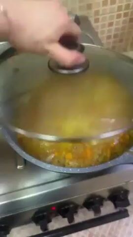 The video shows a pot of soup cooking on a gas stovetop. A hand lifts the glass lid, revealing a yellow broth with chunks of potatoes and other vegetables. Steam rises from the pot. A second shot shows a baked dish covered in foil with a spatula resting on it. The dish appears to have a golden-brown, bubbly surface. The countertop is light-colored and marbled.