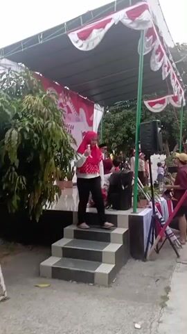 A group of people are gathered outdoors around a small stage decorated with red and white bunting. A woman in a red hijab and a white polka dot shirt is speaking from the stage, holding a microphone. There are speakers and other equipment on the stage. Several people in red shirts are visible in the background, some holding what appear to be flags. The event seems to be taking place in a paved area, possibly a courtyard or sports field, with trees and buildings in the background. The atmosphere appears festive.