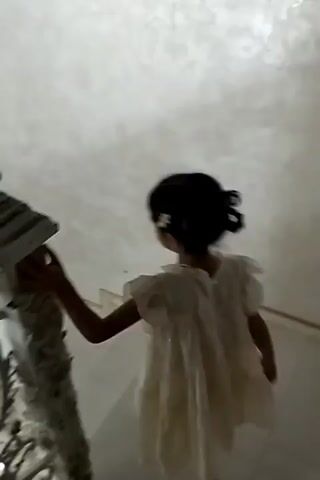 A young girl with dark hair in a bun, wearing a cream-colored dress, is walking up a white staircase. She has a flower tucked behind her ear. The staircase has an ornate white railing with gold accents. To the left of the staircase is a decorative pillar. In the background, blue curtains are drawn. The lighting is dim.