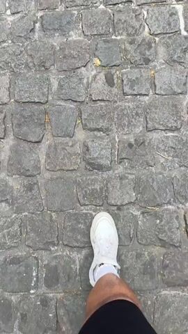 The video opens with a close-up, low-angle shot of a person's right foot, clad in a white sneaker and white sock, stepping on a grey cobblestone street. The perspective then transitions to a wider view, showing the cobblestone path leading towards the grand Notre-Dame de Reims cathedral in the distance. Lush green trees flank the path, framing the impressive Gothic facade of the cathedral.

In front of the cathedral, several people are visible walking and gathering. The sky is overcast, indicating a cloudy daytime setting. Metal barriers and some banners or displays are set up in the square before the cathedral entrance. The camera gradually pans up, centering the cathedral's ornate architecture, including its large rose window and twin towers, within the frame of the surrounding trees. No discernible text is visible on any banners or signs.