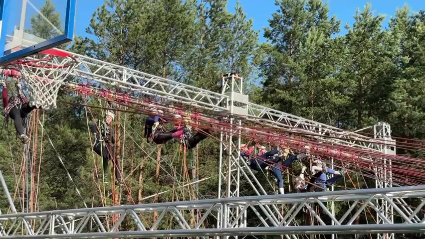 In this media file, a group of people are participating in an outdoor obstacle course. The course appears to be constructed from metal trusses and ropes, with a basketball hoop situated at one end. Several individuals are suspended in the air, utilizing ropes and harnesses to navigate the course. They are dressed in athletic wear, including helmets and protective gear.

The setting is an outdoor environment with a dense forest of pine trees in the background. The sky is clear and blue, suggesting fair weather. The scene is brightly lit, indicating it is daytime, likely late morning or afternoon. The individuals are engaged in various stages of traversing the obstacle course, with some actively climbing and others appearing to rest or prepare for their next move.

The overall atmosphere seems to be one of adventure and physical challenge. The people are focused and determined as they tackle the course. There is no visible text within the media file. The presence of a basketball hoop might suggest a theme or a unique element to this particular obstacle course. The metal structure is large and complex, indicating a professionally designed and installed course. The use of ropes and harnesses highlights the element of height and risk involved in the activity.