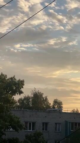 A short video captures a view of the sky above a building. The sky is filled with white and gray clouds, illuminated by the soft light of what appears to be sunrise or sunset. Two black wires are visible diagonally across the frame, obscuring parts of the sky. Below the sky, the top of a multi-story building is visible, with a line of trees peeking over the roof. The building has multiple windows and appears to be made of brick or a similar material. The overall scene is calm and serene, with the natural beauty of the sky contrasting with the man-made structures.