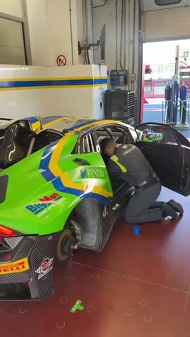 A person wearing a black shirt and grey pants is kneeling beside a green and yellow race car in a garage. The car has the word "PIRELLI" on the back tire and "FENERI" on the side. The person appears to be working on the car's interior or door. Another person in a black t-shirt walks by in the background and waves. There are other race cars visible in the garage, as well as equipment and tools. The garage appears to be a professional racing facility.
