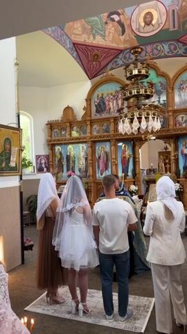 The video shows an indoor scene, likely a church. In the foreground, from right to left, are four people facing away from the camera. The person on the far right wears a white suit and headscarf. Next to them is a woman in a white wedding dress and veil, with a man in a white t-shirt and jeans standing to her left. Another woman, in a brown dress and headscarf, is to the left of the bride. A clergyman, wearing a blue and gold robe, stands in front of the couple, facing them. The background features an ornate iconostasis, typical of Orthodox churches, with religious icons and murals on the walls and ceiling. A chandelier hangs from the ceiling. Candles are visible in the lower corners of the frame. The people appear to be participating in a religious ceremony. The audio is in Romanian, with the clergyman speaking.
