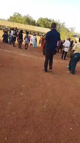 The video shows a small crowd of people standing in a dirt lot. There is a man in a navy shirt and black pants walking through the lot. The people are wearing a variety of clothing, including traditional African prints. Some of the people are talking to each other. In the background, there is a long building with a metal roof. The sky is clear and blue.