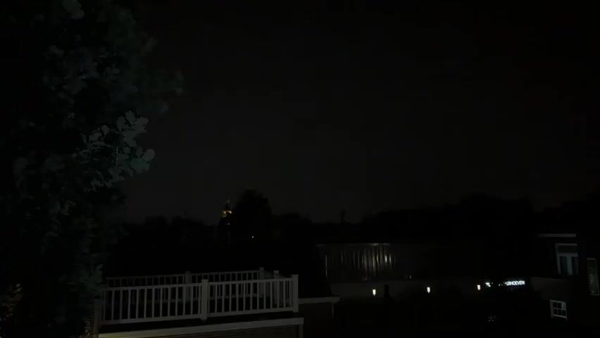 The video shows a nighttime outdoor scene. In the foreground, a portion of a tree is visible on the left side of the frame. Below the tree and extending across the lower part of the image is a white railing, likely part of a deck or balcony. Beyond the railing, several dark silhouetted structures, presumably houses or buildings, are visible. In the distance, silhouetted trees and a structure that appears to be a church steeple are faintly discernible against a dark, cloudy sky. The overall lighting is dim, with occasional faint illumination in the sky, suggesting it might be overcast or there could be distant light sources. There are no people or active events depicted in the video.