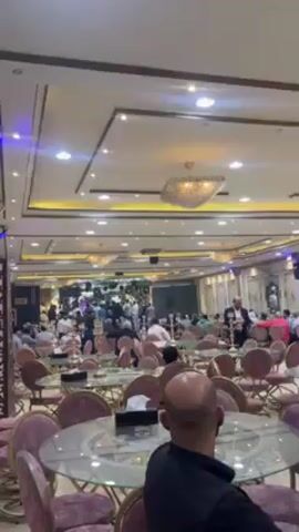 The video shows the interior of a large banquet hall filled with people. The hall is decorated with elegant chandeliers and ornate ceiling details. Round tables with chairs are arranged throughout the space. Many of the people are standing near a raised stage area, where there appears to be a performance or gathering taking place. Some people are standing at the tables, possibly talking or observing the event on the stage. The atmosphere suggests a celebratory event or large social gathering.