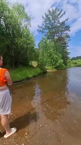 This media file captures a serene riverside scene, likely in Dachnaya, Russia. The primary subjects are the natural landscape and a body of water. A calm, clear river flows gently, with a sandy, pebbly bottom visible in the foreground. Lush green trees and vegetation line the riverbanks, creating a verdant and inviting atmosphere. Tall pine trees stand prominently among the foliage, reaching towards a bright blue sky dotted with fluffy white clouds.

The activity taking place is fishing. A fishing rod is visible extending into the water from the left side of the frame, indicating someone is actively engaged in the sport. In the distance, a dark-colored vehicle, possibly an SUV, is parked near the riverbank, with a few people gathered around it. The presence of the vehicle and people suggests a recreational outing.

Notable details include the clear, sunny weather, indicated by the bright blue sky and the sunlight reflecting off the water. The time of day appears to be midday or early afternoon, given the quality of the light. The location cues, such as the type of trees and the overall natural scenery, are consistent with a rural or wilderness setting in Russia. The overall mood conveyed is one of peace, tranquility, and enjoyment of nature. There is no visible text in the media file.