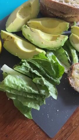 The video shows a close-up, overhead shot of food being prepared on a dark gray cutting board placed on a wooden surface. Sliced avocados, romaine lettuce, and toasted bread with butter are visible. In the upper left corner, a whole lemon is present. The actions captured include slicing and dicing, with a hand briefly entering the frame to sprinkle something from a shaker, likely salt or pepper, onto the lettuce. The lighting is natural and comes from above and to the right, casting soft shadows. The background is slightly blurred, with hints of a blue surface in the upper left and the wooden table.