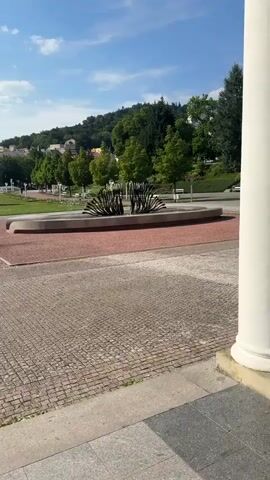 The video shows a scenic view of a park on a sunny day. The foreground is paved with cobblestones, leading to a pathway with a reddish-pink surface. In the middle ground, there is a modern sculpture that resembles a stylized plant or fountain. Beyond the sculpture, a well-maintained green lawn stretches out, dotted with lampposts.

In the background, a lush green hillside is covered with trees and dotted with buildings, suggesting a town or village nestled within the landscape. The sky is bright blue with scattered white clouds, indicating fair weather and likely daytime.

There are no people clearly visible in the video, though there might be distant figures on the pathways. The overall impression is one of tranquility and natural beauty, characteristic of a park or garden. The architecture of the distant buildings and the style of the park's landscaping suggest a European setting, and the provided context confirms this as Mariánské Lázně, Czechia. The lighting indicates it's likely late morning or early afternoon. No text is visible in the media.