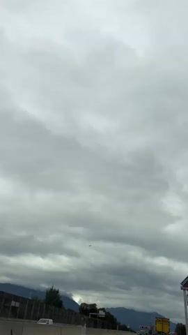 The video captures a view of a cloudy sky, likely from a moving vehicle. A small airplane is visible in the distance, flying through the clouds. In the foreground, there are elements that suggest a road or highway, including a barrier and road signs. One sign in particular reads "Salzburg Designer Outlet." Trees and distant mountains can also be seen along the horizon. The overall atmosphere is overcast and somewhat gray, with the clouds dominating the frame.
