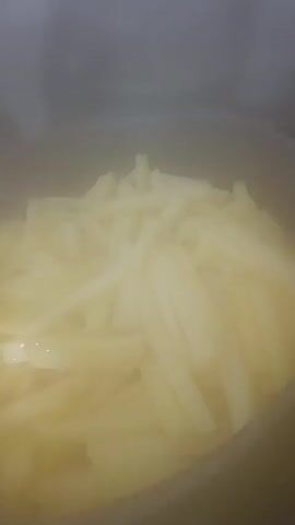 The video shows a close-up view of thinly sliced potatoes in a pot on a stovetop. Steam is rising from the pot, indicating that the potatoes are cooking. The pot appears to be dark-colored and has a speckled pattern. The lighting in the video is dim, and the focus is on the potatoes. The video also shows a hand briefly lifting a glass lid off the pot, allowing more steam to escape. The background is out of focus and appears to be a kitchen environment.