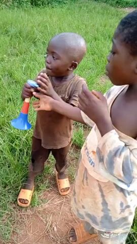 The video shows four young Black children standing in a grassy field. Three of the children are playing with plastic toy horns. One child is holding a horn with a blue bell, while another is playing a red and yellow toy horn. The third child is standing to the right, looking at the others. The children are wearing casual clothes, and two of them have sandals. The background is green with trees and foliage visible.