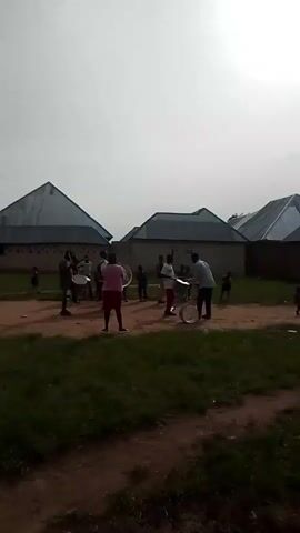 This media file captures a group of people, primarily children and young adults, engaged in what appears to be a musical rehearsal or practice. The subjects are gathered outdoors in a dirt and grass area, with several houses featuring distinctive, slanted metal roofs in the background, indicative of a location in Bokkos, Nigeria.

The central activity involves a drum ensemble. Several individuals are holding and playing drums, including what looks like a bass drum positioned in front of a person wearing a pink sweater and a red skirt. Another person in a white shirt and red pants is holding a smaller drum. Other members of the group are standing around, some holding drums and mallets, suggesting they are either waiting for their turn or participating in a marching band or similar musical activity. The presence of multiple drums, some large and some smaller, implies a structured musical performance is being prepared.

The time of day appears to be daytime, with the sky visible in the background being overcast or hazy, suggesting potentially cloudy weather. The overall atmosphere seems casual and participatory, with the individuals focused on their musical task. There are no overtly strong emotional cues visible, but the engagement with the instruments suggests a sense of purpose and perhaps enjoyment.

The visible text on the large bass drum appears to read "YAMAHA," which is a well-known manufacturer of musical instruments. This reinforces the context of a musical activity. The setting is modest, with unpaved ground and simple structures, fitting the context of a community gathering for a musical purpose in Nigeria.