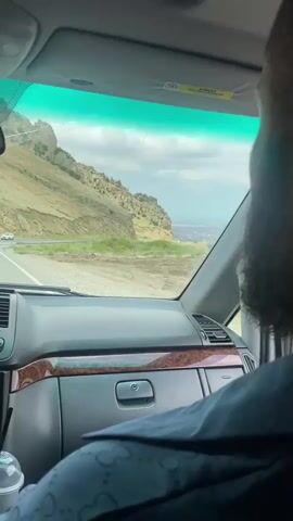The video shows the interior of a vehicle from the passenger's perspective. A man with a beard is seated in the passenger seat, looking out the window. Outside the window, a road curves around a rocky, mountainous landscape under a partly cloudy sky. The dashboard of the vehicle is visible, including a multimedia screen and air vents. A cup with a straw is in a holder on the dashboard. There are air fresheners hanging from the rearview mirror. The vehicle appears to be in motion.