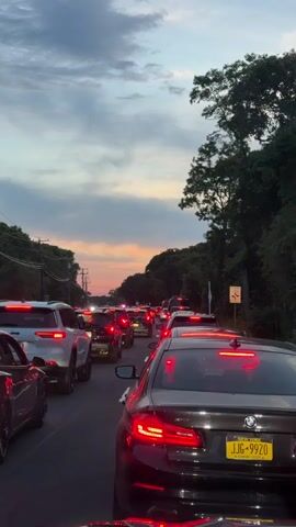 A vertical, eye-level, medium shot captures a traffic jam at dusk. In the foreground, a black BMW sedan is visible from the rear. Its taillights are illuminated, and a yellow license plate reading JJG-9920 is attached. To the left of the BMW, the side of a silver car is visible. In the mid-ground, a white Jeep SUV is seen from the rear, with its taillights illuminated. Numerous other vehicles are lined up behind it, their red taillights glowing. The road is bordered by trees and utility poles on both sides. The sky above is filled with clouds, and a sunset can be seen in the distance, casting an orange and pink glow. A traffic sign is visible on the right side of the road.
