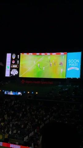The video shows a soccer game being played at night in front of a large crowd. The stadium is lit by bright overhead lights. A large scoreboard displays the score as 1-1. The players are wearing white and dark-colored uniforms, and they are spread across the field. The stands are full of spectators, many wearing white. There are advertisements visible on the sidelines and on the upper deck of the stadium. A person holding the camera moves it to show different views of the game and crowd.