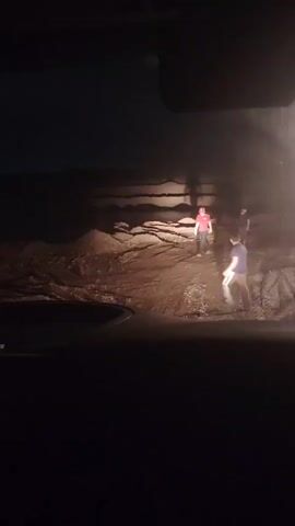 This video captures a group of four individuals standing on a dirt road at night. A bright light, possibly from a vehicle's headlights, illuminates the scene from the right side of the frame, casting long shadows. The individuals appear to be engaged in conversation or observation. The environment seems to be a somewhat uneven terrain with piles of dirt or rubble. The overall impression is one of a brief, unscripted encounter in a dimly lit outdoor setting.