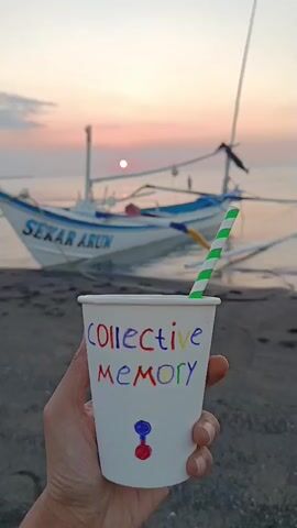 A hand holds a white paper cup with the words "COLLECTIVE MEMORY" written on it in colorful letters. Below the text are two small, colorful circular shapes, one red and one blue, stacked vertically. A green and white striped straw is inserted into the cup. The background shows a calm body of water with a white and blue boat docked on a sandy or dark-colored beach. The sky above is a soft gradient of pink and orange, indicating either sunrise or sunset, with a small, dim sun visible. The overall scene is peaceful and serene.