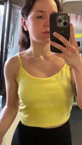 A woman with curly dark brown hair, wearing a yellow spaghetti-strap top and black leggings, is taking a selfie video in what appears to be a home gym. The woman is holding her phone in her right hand, partially obscuring her face. She is facing a mirror, and a window is visible to her left. A weight machine is behind her. She is sweating, and a small plastic cup is on the windowsill. She turns slightly to the left and then back to the front while holding the camera. She then puts her hair up.